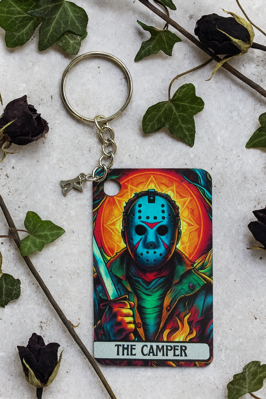 Keychain with 'The Camper' design on a textured surface with leaves and flowers.